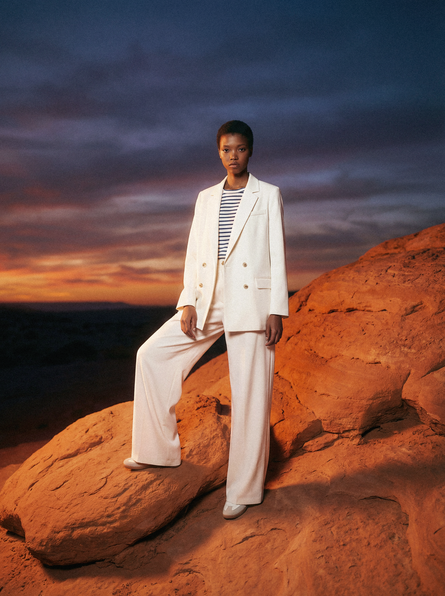Fashion model in white suit poses against dramatic red rock formations at sunset creating striking desert contrast.