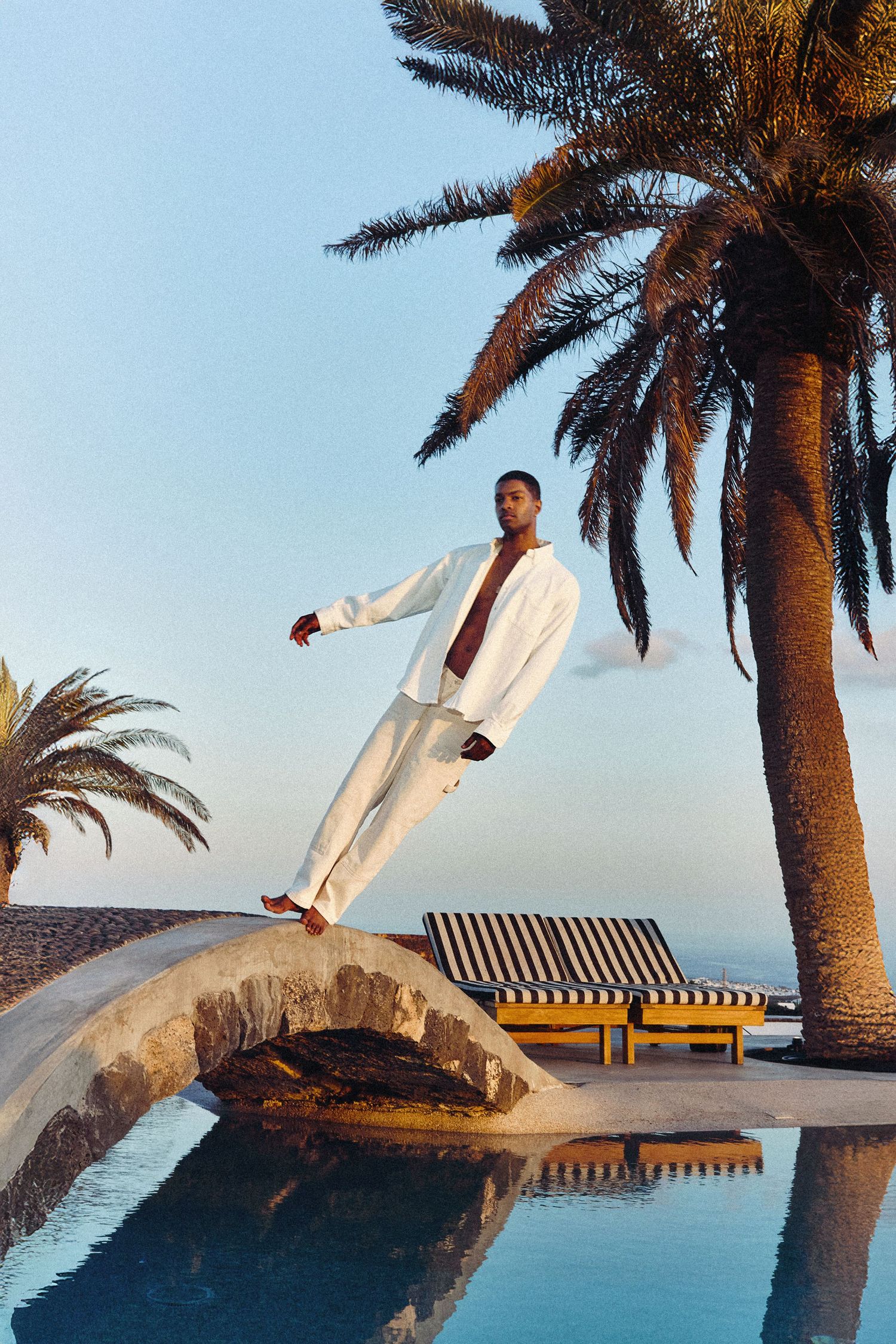Figure in white clothing poses on curved stone structure by luxurious poolside with palm trees at sunset.