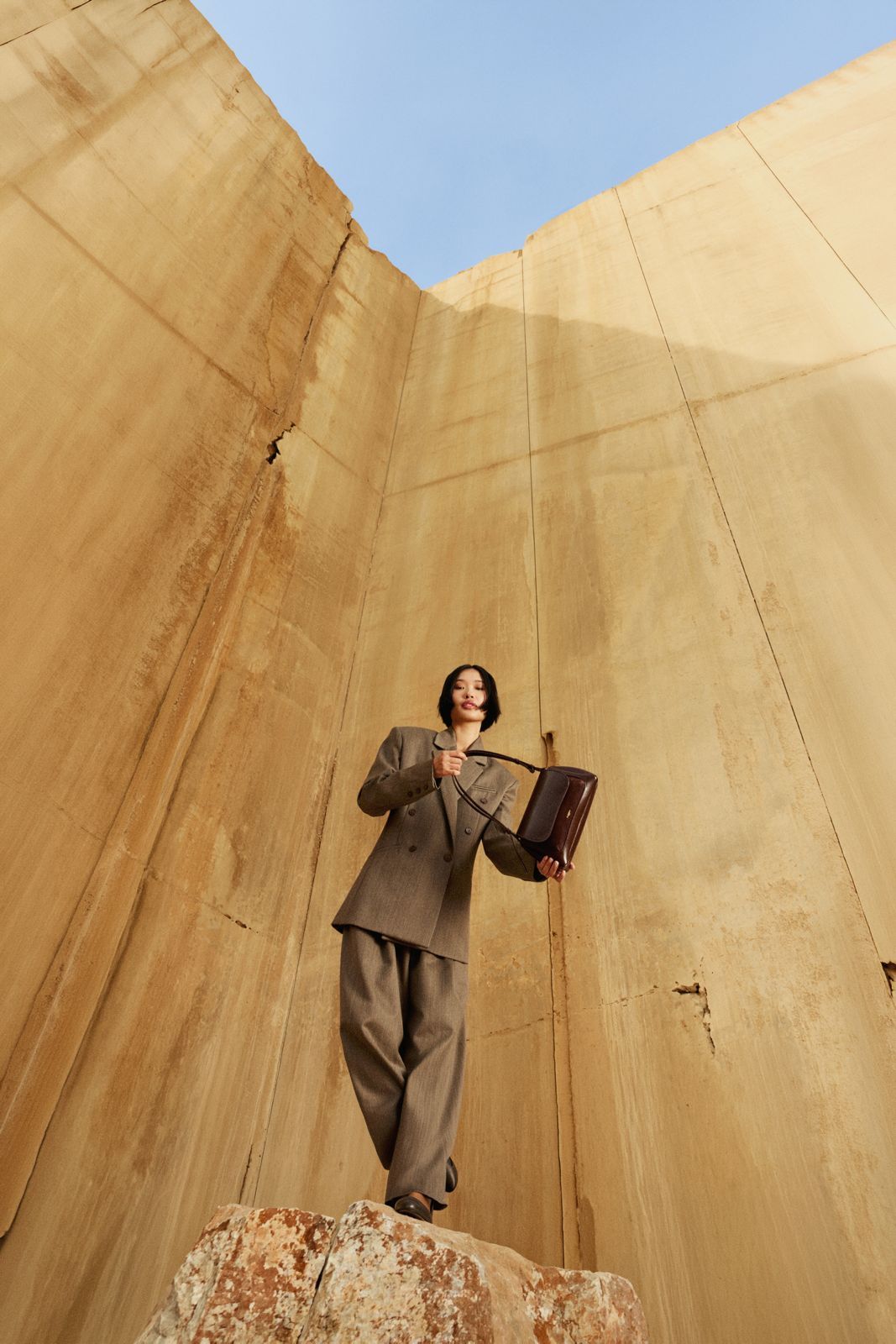 A person in a gray suit holds a briefcase while standing on a rock against a large sandy wall under a blue sky.