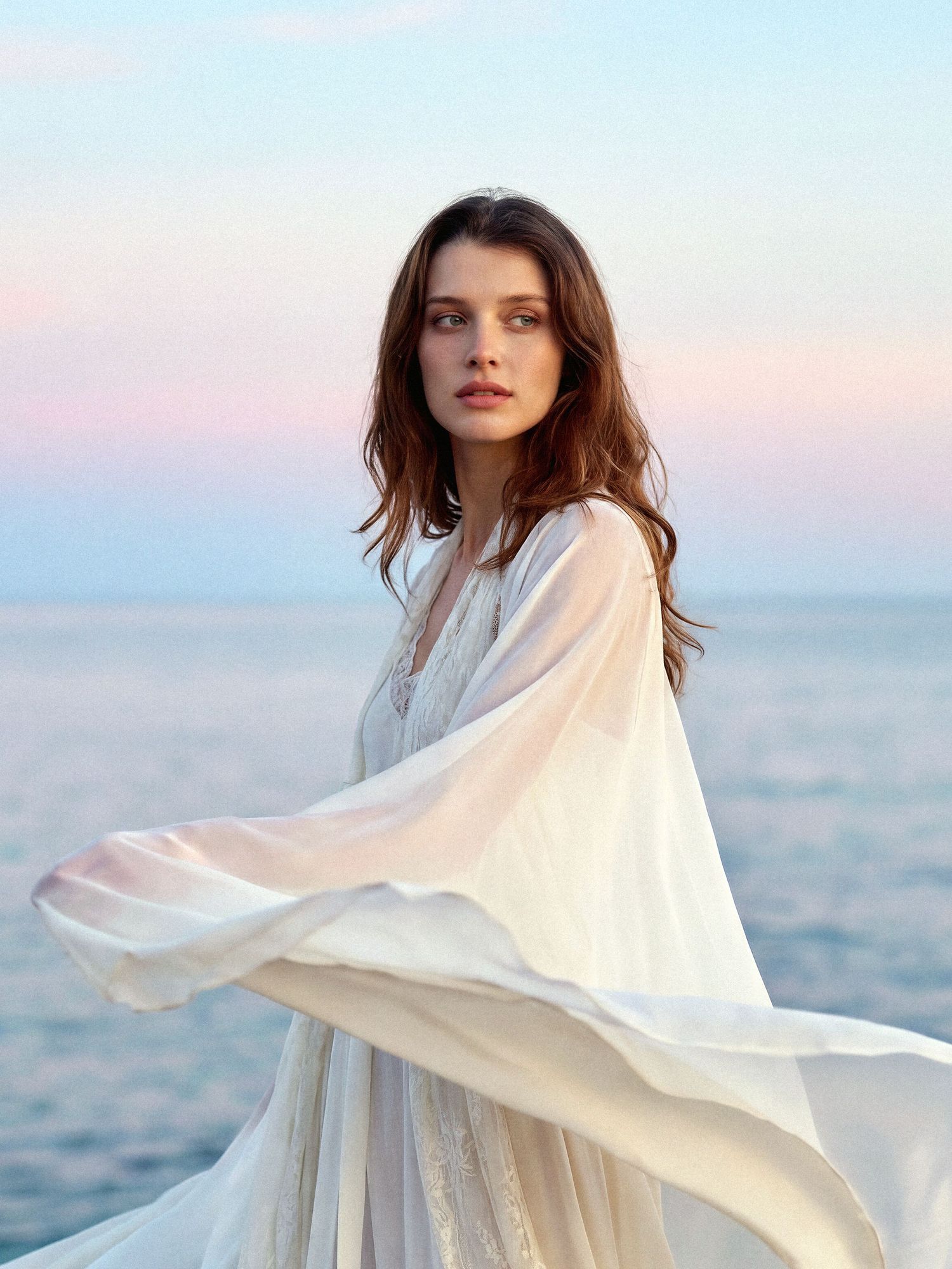 A model in a flowing white dress poses gracefully against a pastel sunset sky over the ocean.