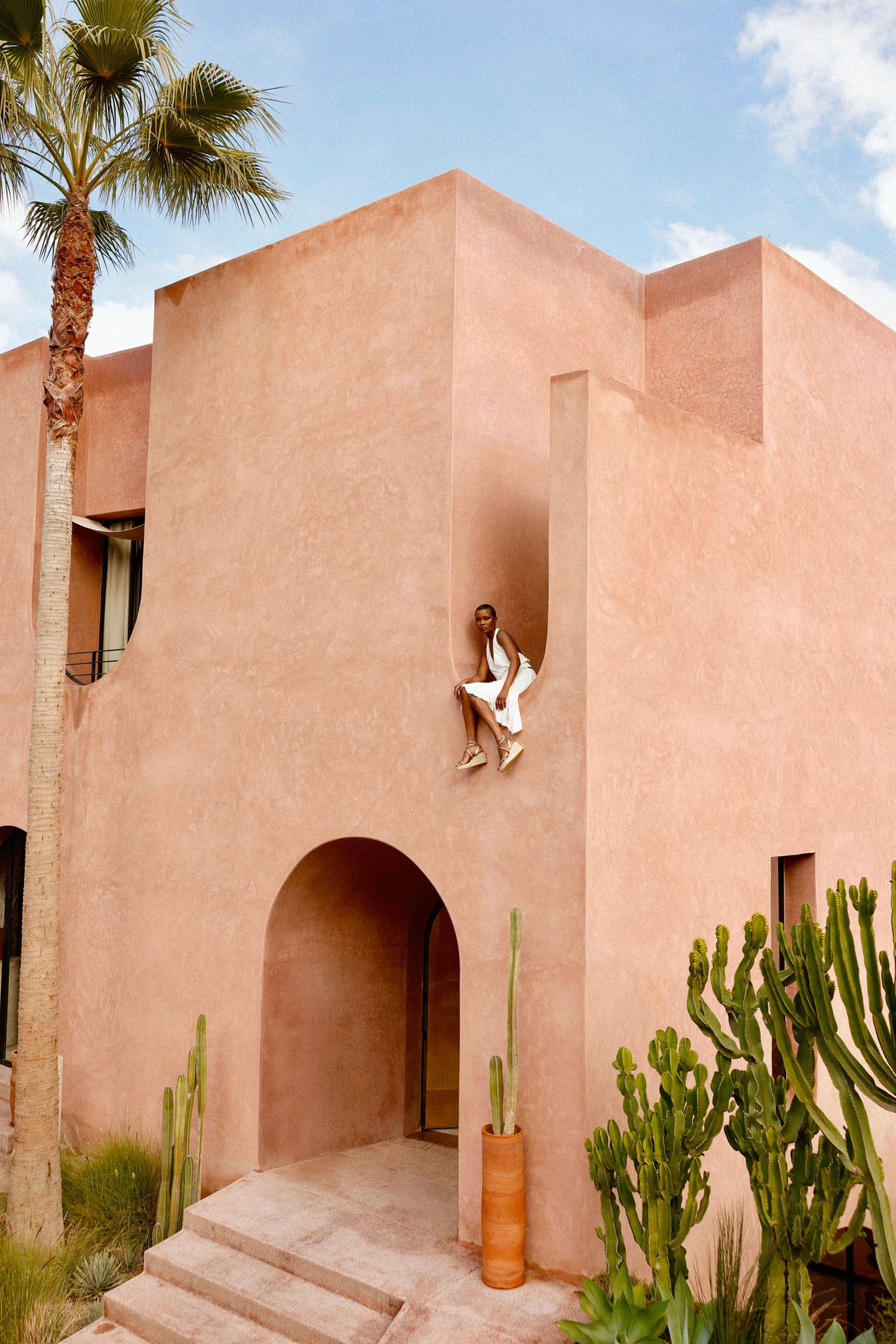 Modern pink stucco building with arched entrance, desert landscaping and palm trees against blue sky.