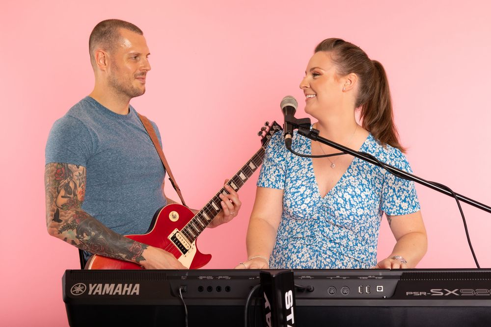 Musicians performing together with guitar and keyboard against pink background.