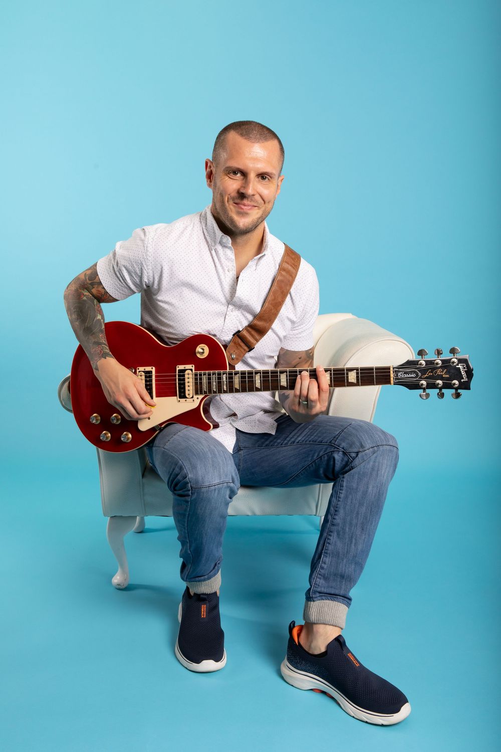 Musician in white shirt sitting with red electric guitar against blue backdrop.
