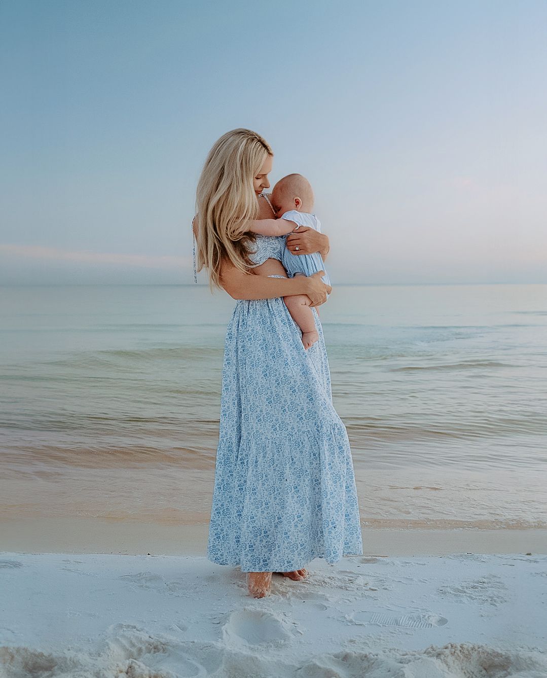 A tender moment captured at the beach with a parent and baby in matching light blue outfits against the ocean backdrop.