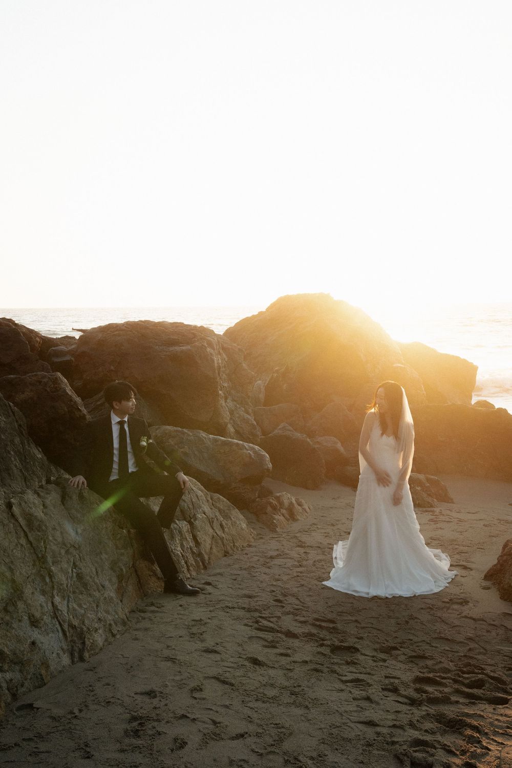 A bride in a white wedding dress stands on a rocky beach at sunset while golden light illuminates the scene.