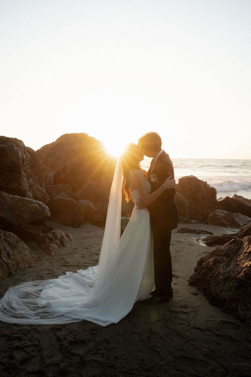 Romantic beach wedding scene at golden hour with couple embracing among coastal rocks and crashing waves.