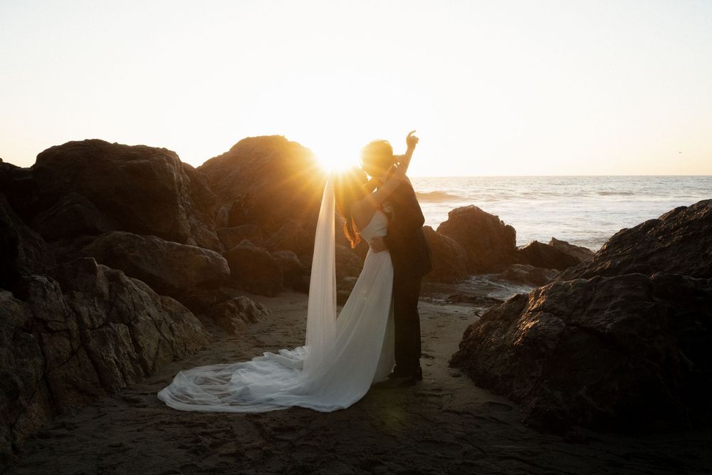 Silhouetted figures in wedding attire stand on rocky coastal cliffs at sunset with flowing white dress in the breeze.