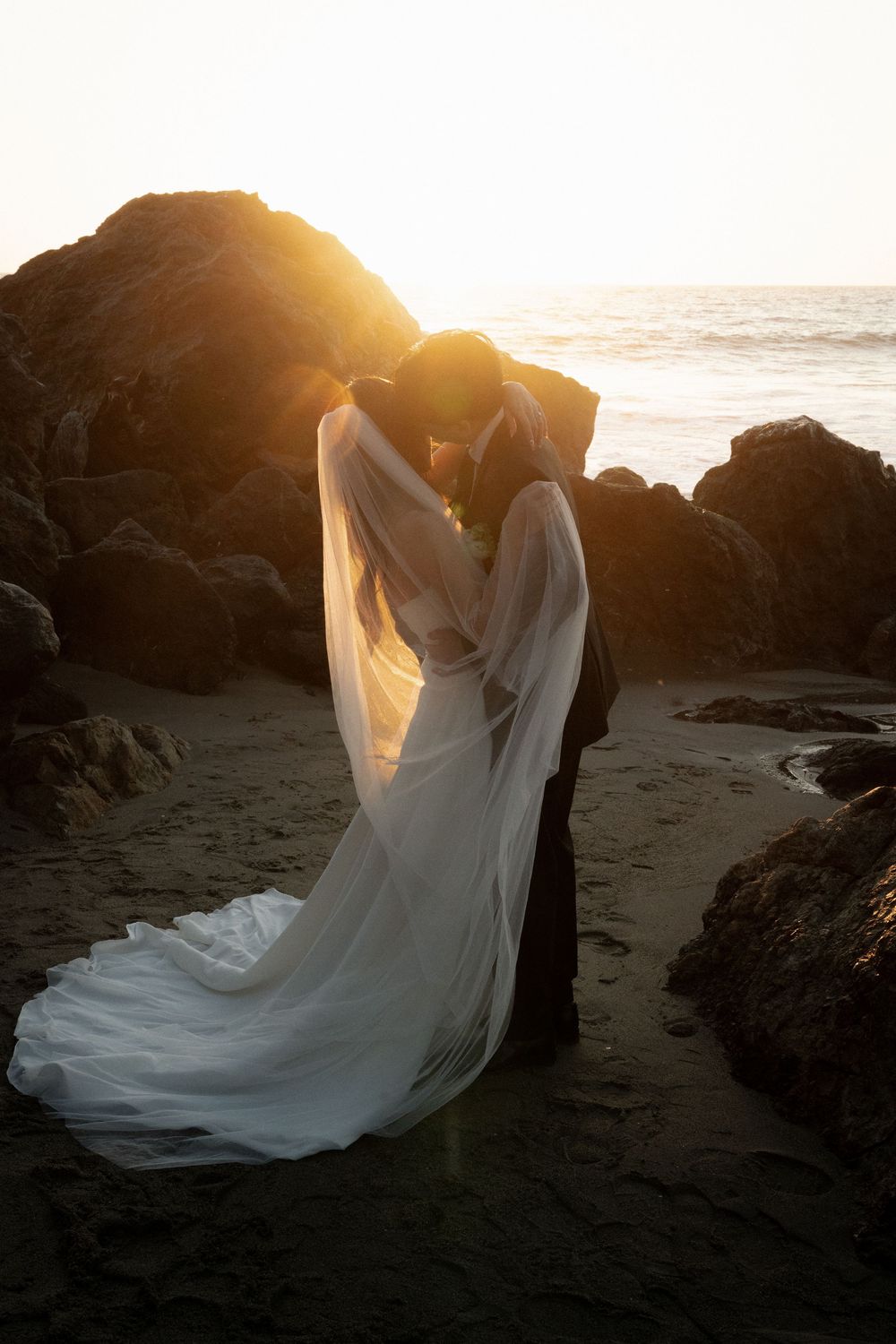 A flowing white wedding dress billows in the golden sunset light against dark coastal rocks on a beach.