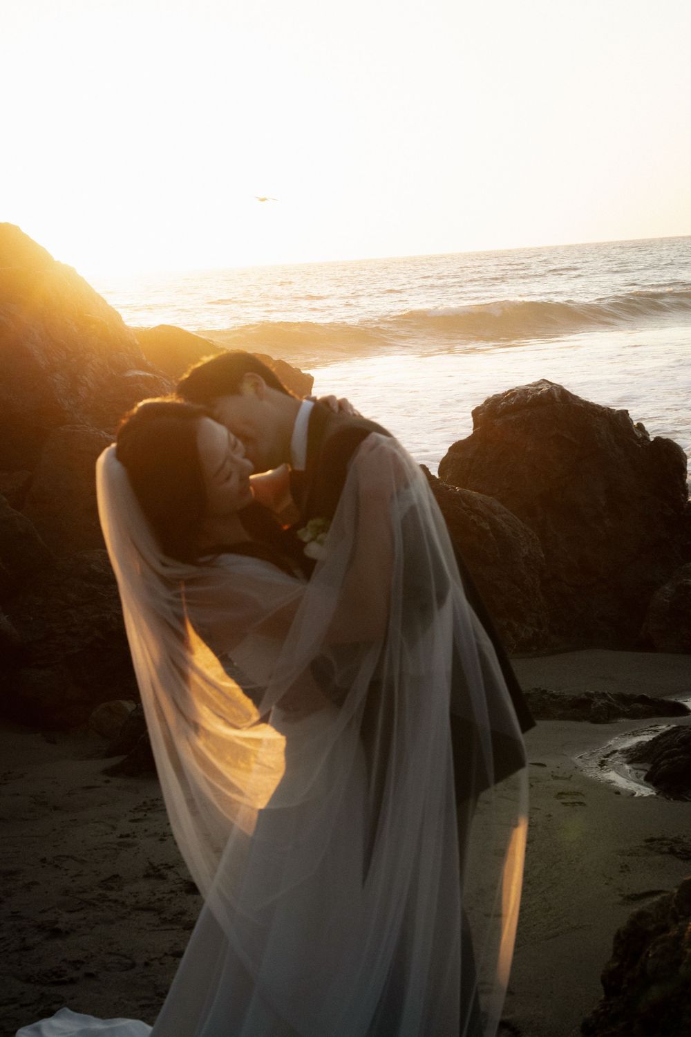 A figure in a flowing white dress stands against a dramatic sunset on a rocky beach coastline.