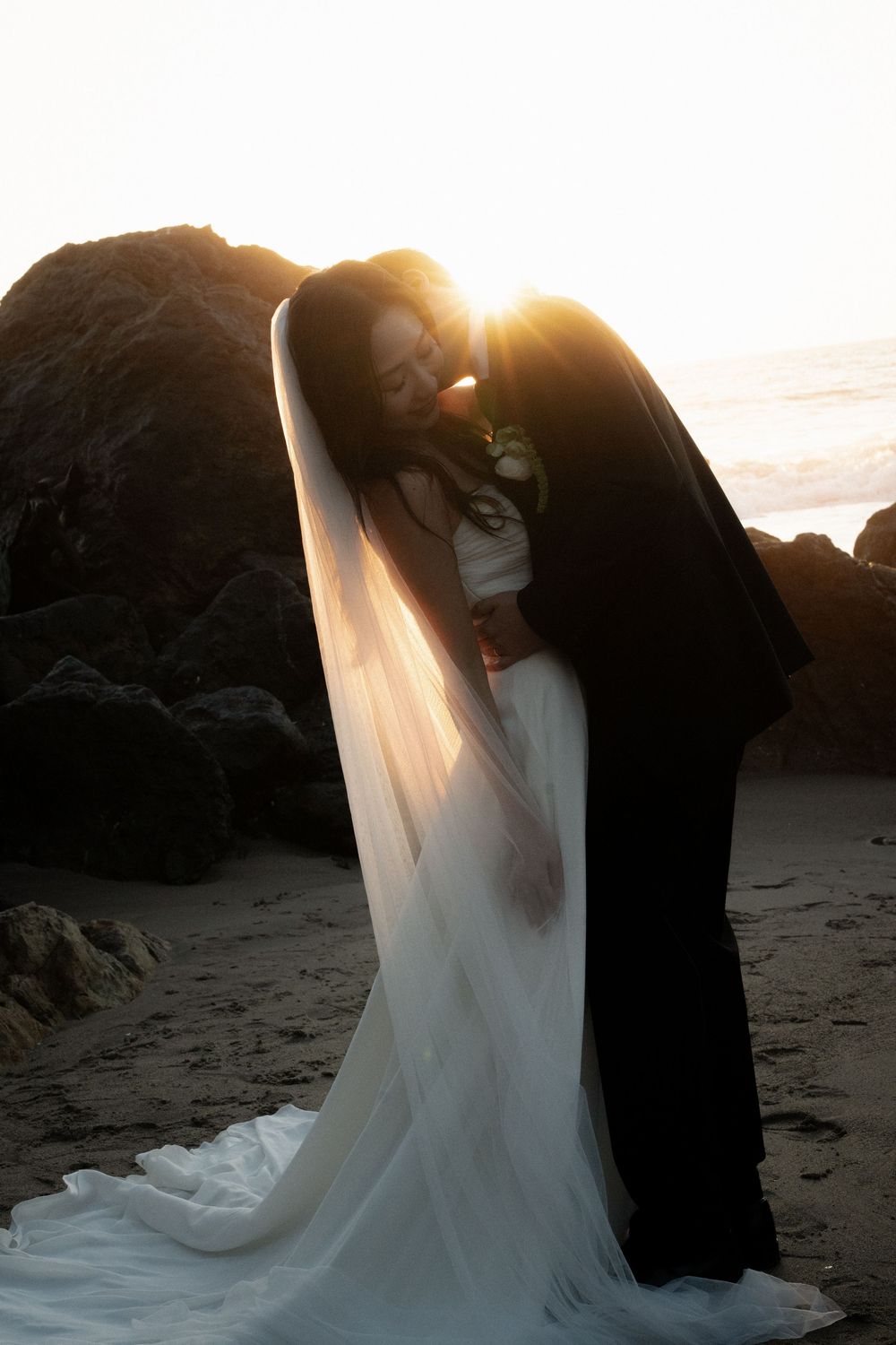 A romantic sunset beach scene with a flowing white wedding dress silhouetted against rocky cliffs.