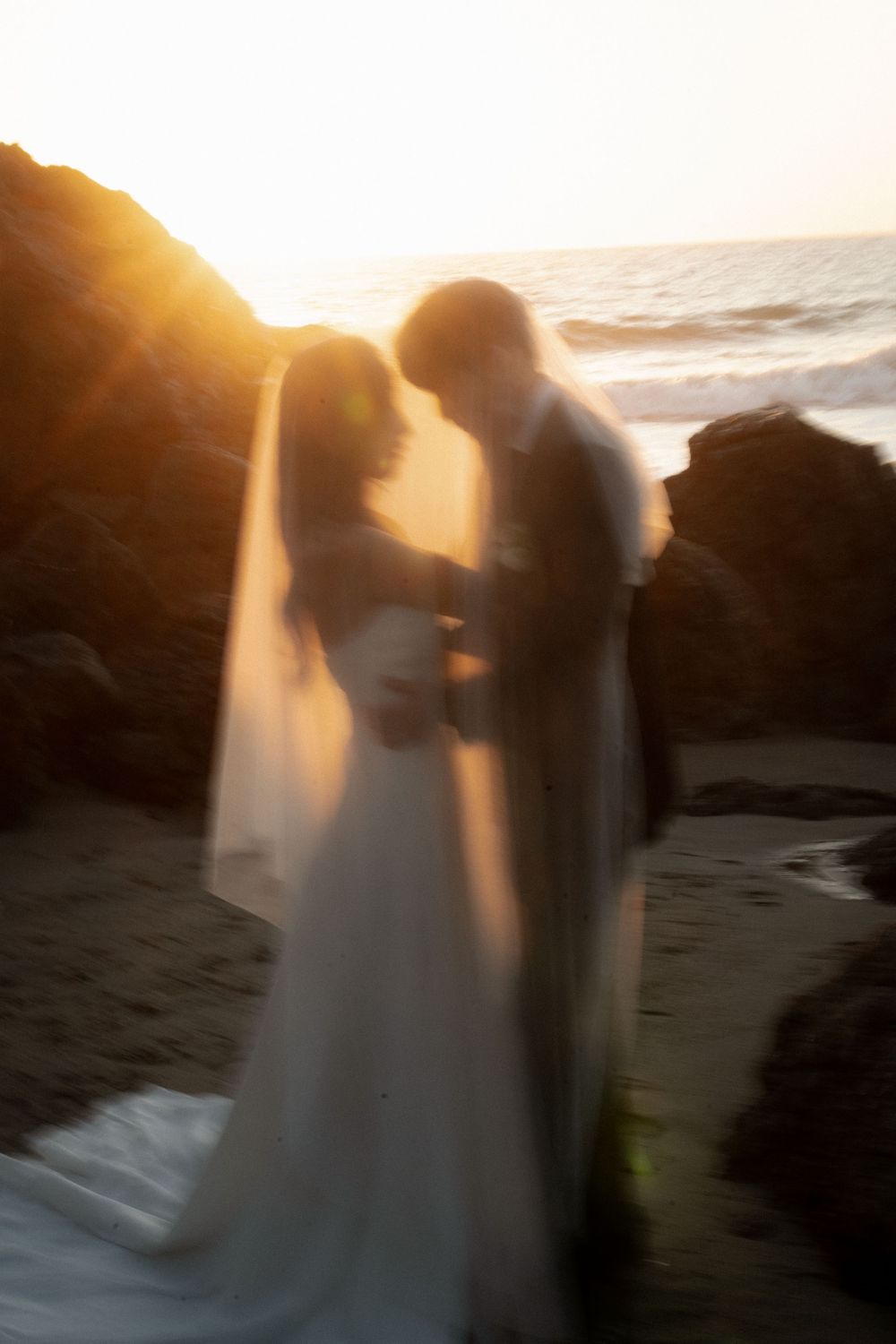 Romantic sunset silhouette of a couple on a beach with golden light streaming through a flowing white veil.