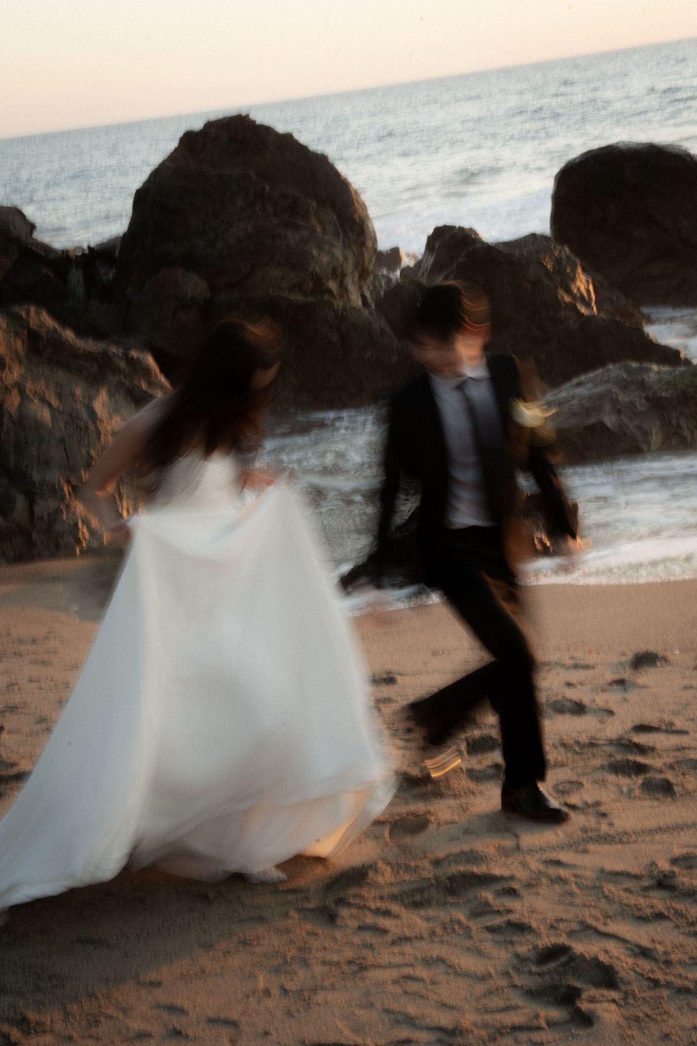 Motion-blurred figures dance on a rocky beach at sunset, creating a dreamy and romantic seaside atmosphere.