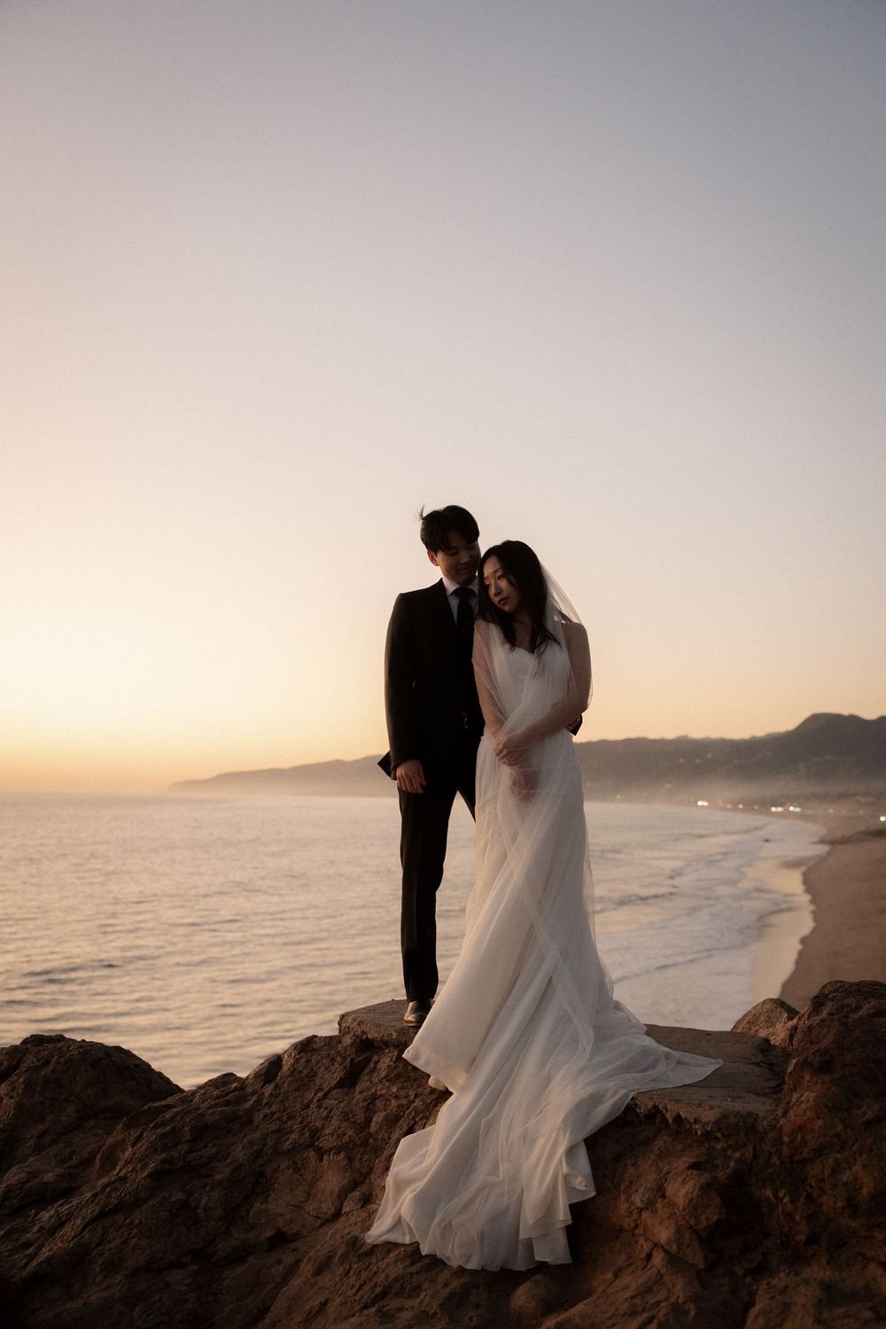 A couple in wedding attire poses together on coastal rocks during golden hour at the beach.