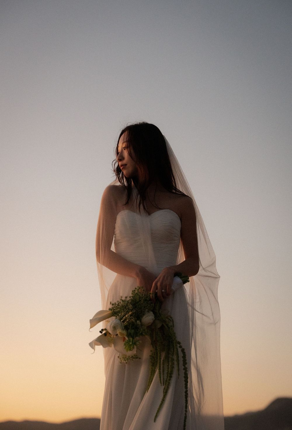 Ethereal bridal portrait silhouetted against a soft sunset sky with delicate veil movement.