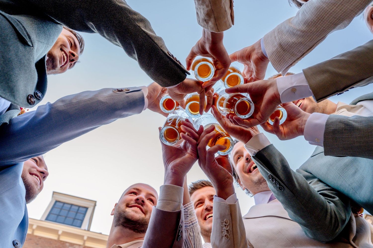 Group of friends toasting with beer bottles and glasses in a circle from below perspective.
