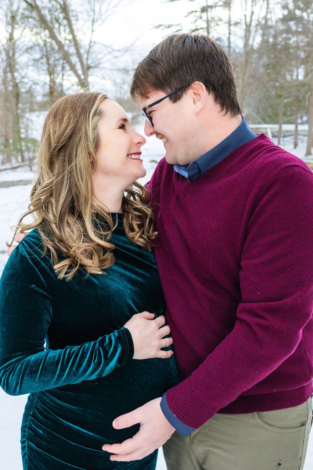 A smiling pregnant couple embraces and gazes at each other in a snowy winter maternity portrait outdoors.