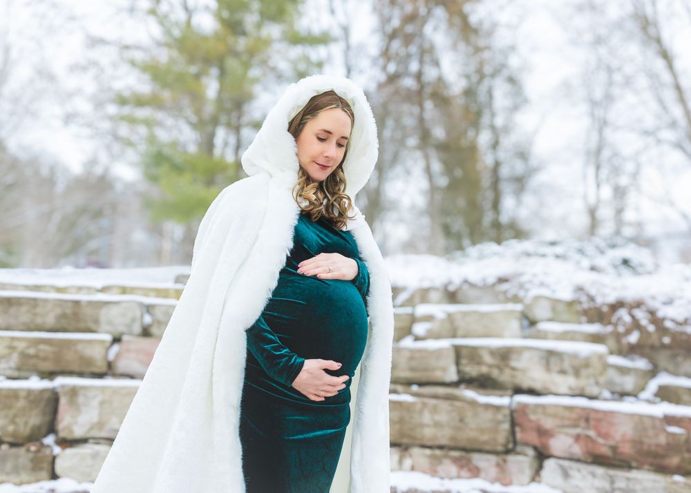 A pregnant woman in a teal velvet gown and white hooded cape cradles her bump in a snowy winter setting.