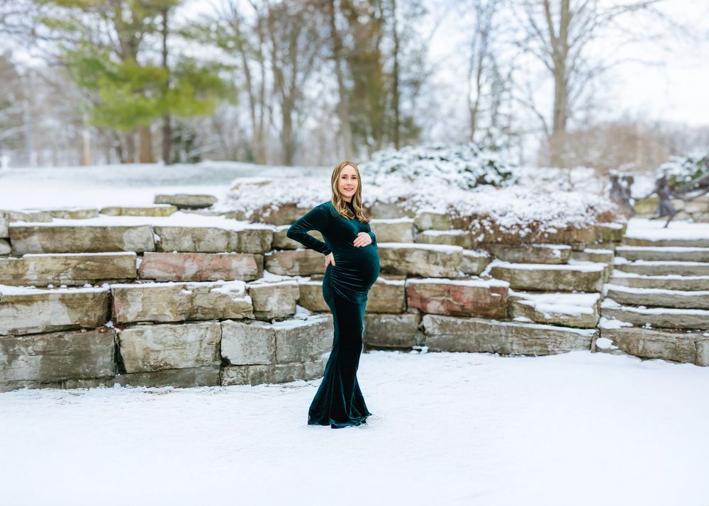 A pregnant woman in a form-fitting dark green velvet gown poses elegantly in a snowy outdoor landscape.