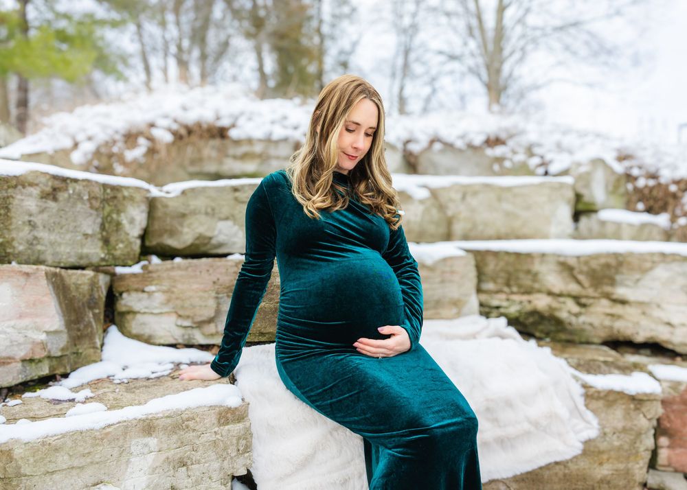 A pregnant woman in a teal velvet gown sits on stone steps cradling her baby bump in a snowy setting.