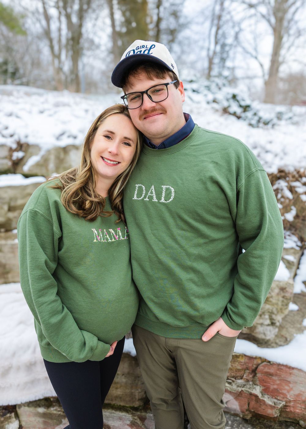 Couple wearing matching green Mama and Dad sweatshirts posing outdoors in snowy winter setting for pregnancy announcement photo.