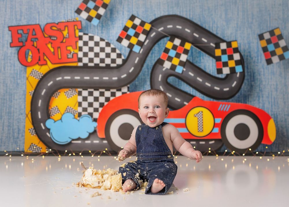 First birthday cake smash photo session with racing car theme backdrop and decorations on blue background.