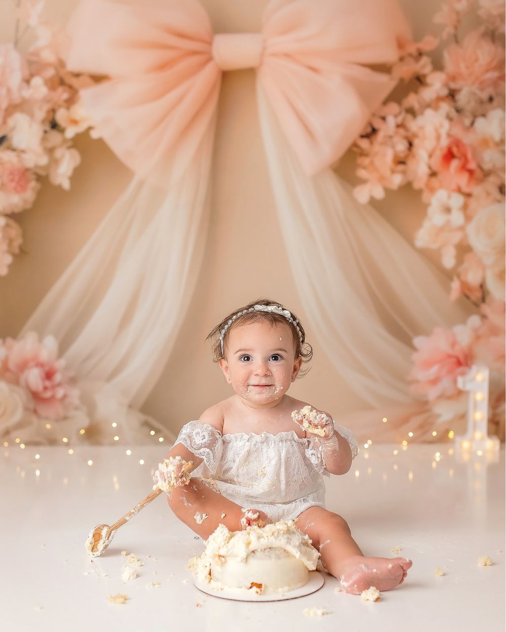 A cake smash photo session featuring a baby in white lace dress with pink tulle decorations and string lights.