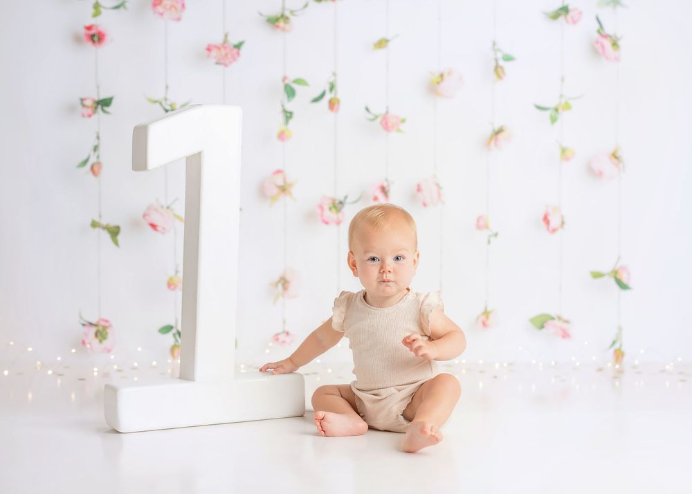 Baby in grey onesie sits and smiles against floral wallpaper background for first birthday photo session.