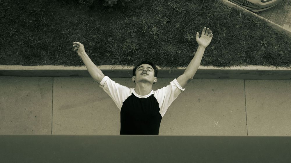 Young man with arms raised looking up toward the sky in a black and white outdoor photo.