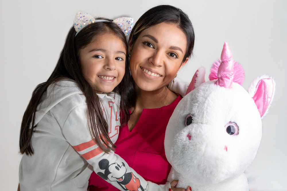 Smiling mother and young daughter posing together with a large white unicorn stuffed animal toy.