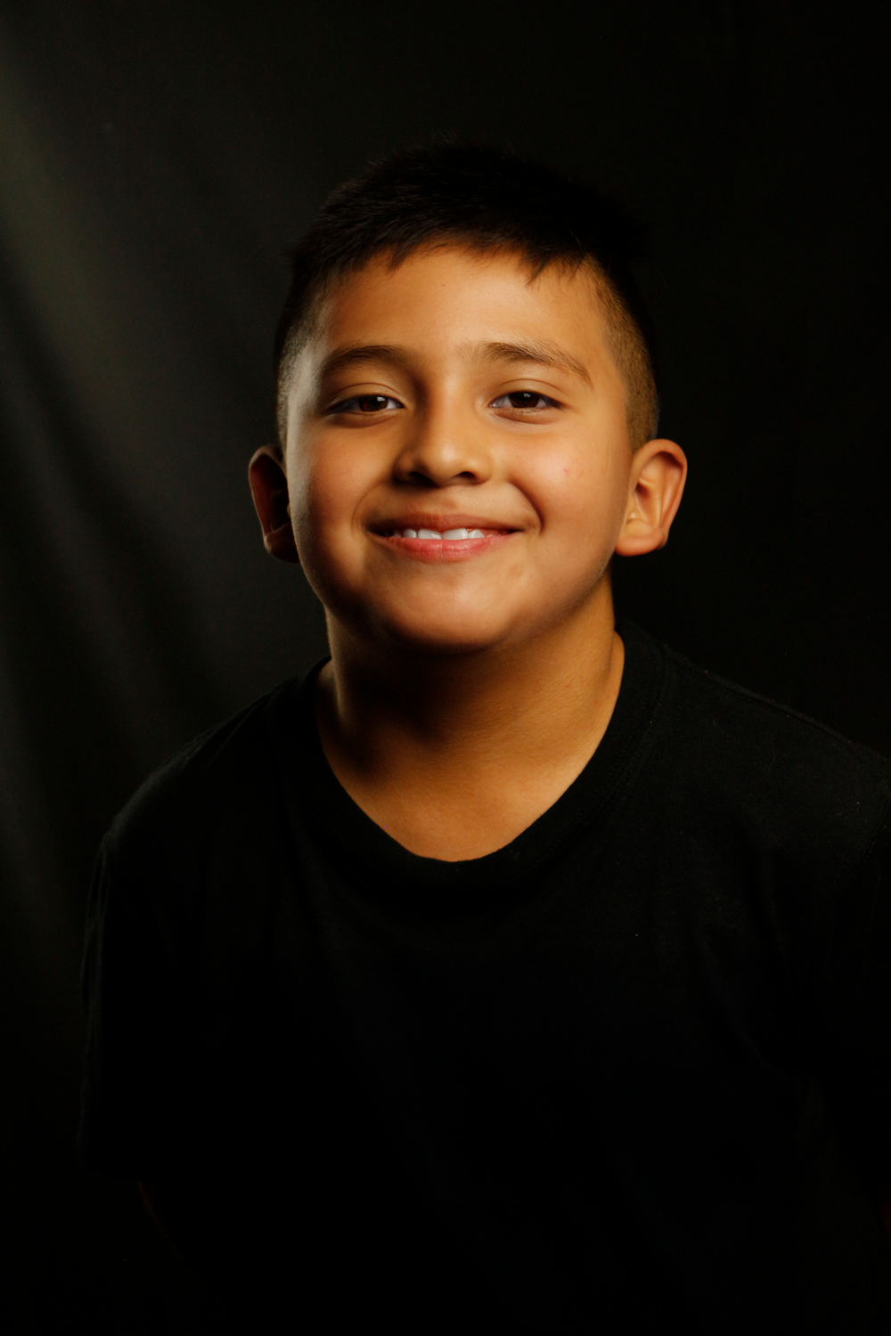 Smiling young Latino boy in black shirt posing for studio portrait against dark background.
