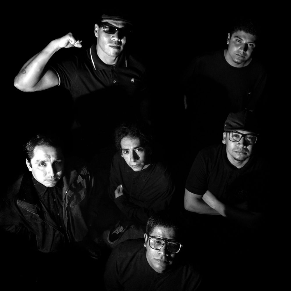 Black and white group portrait of six musicians posing dramatically in dark moody lighting.