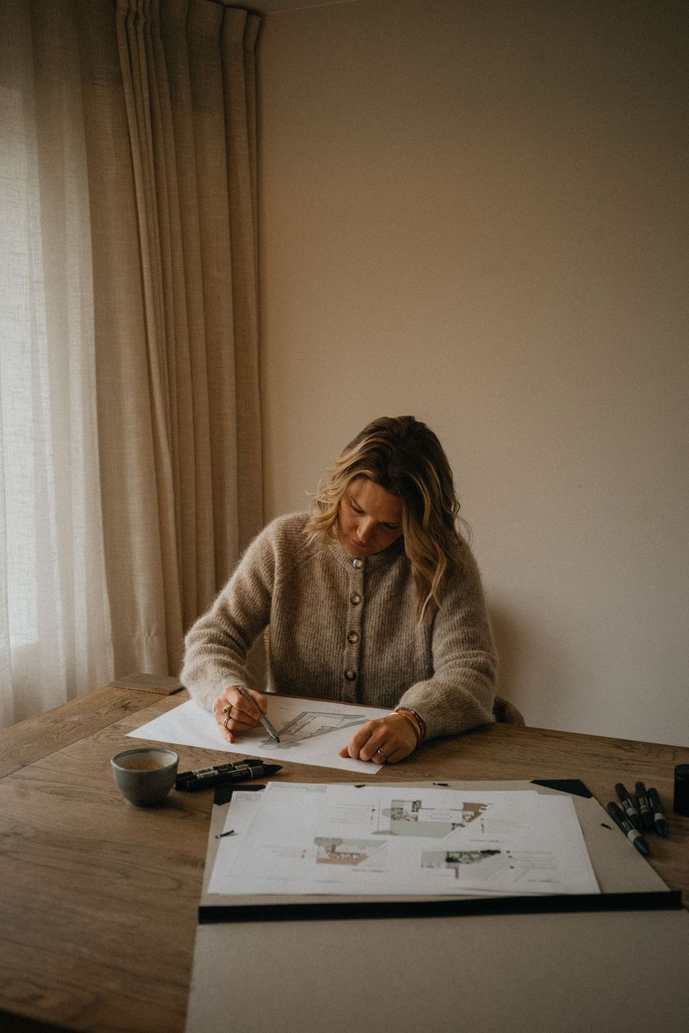 A person in a gray sweater sits at a table working on sketches and drawings near a window with natural light.