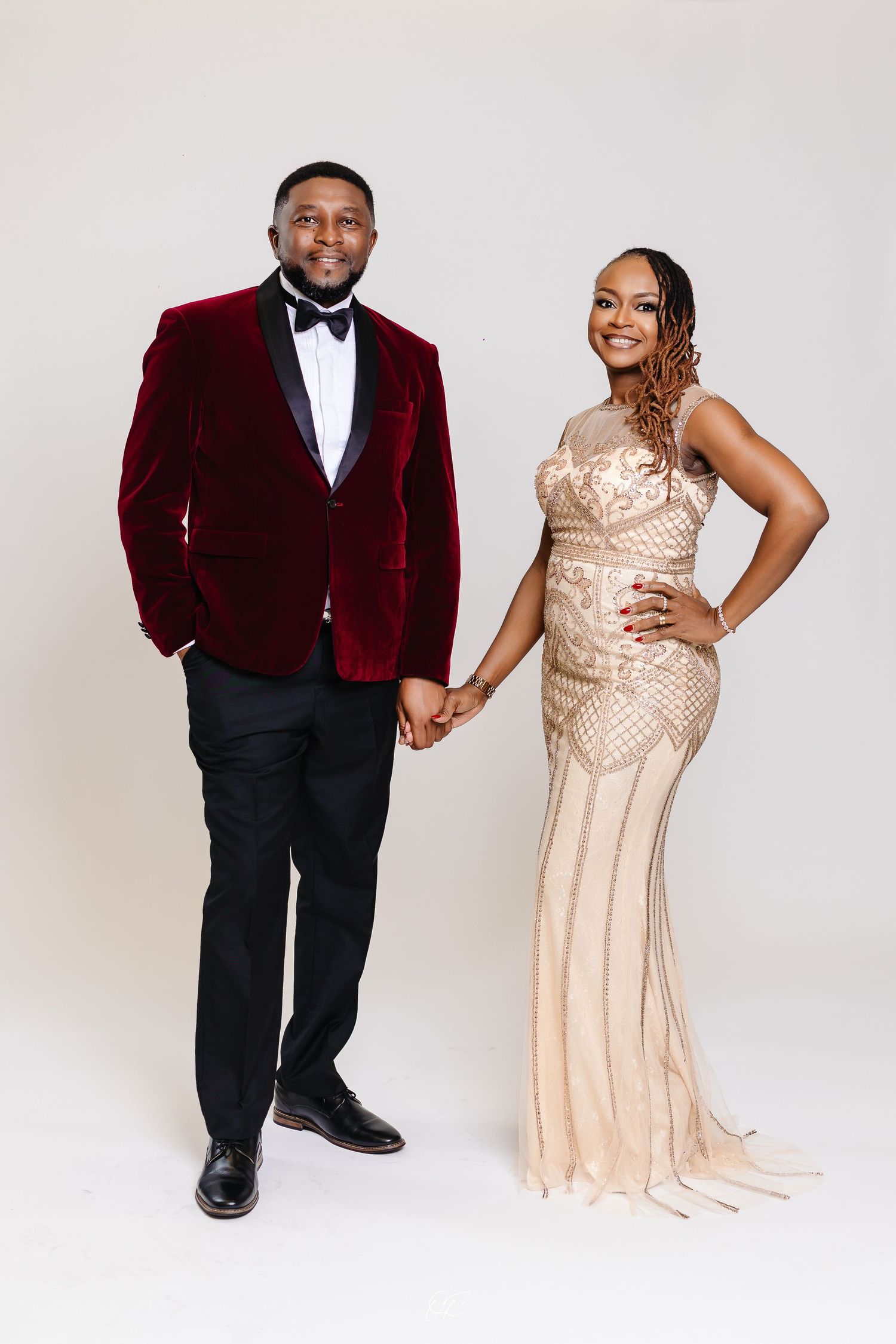 Elegantly dressed couple poses together against white backdrop, wearing burgundy velvet tuxedo and champagne sequined gown.