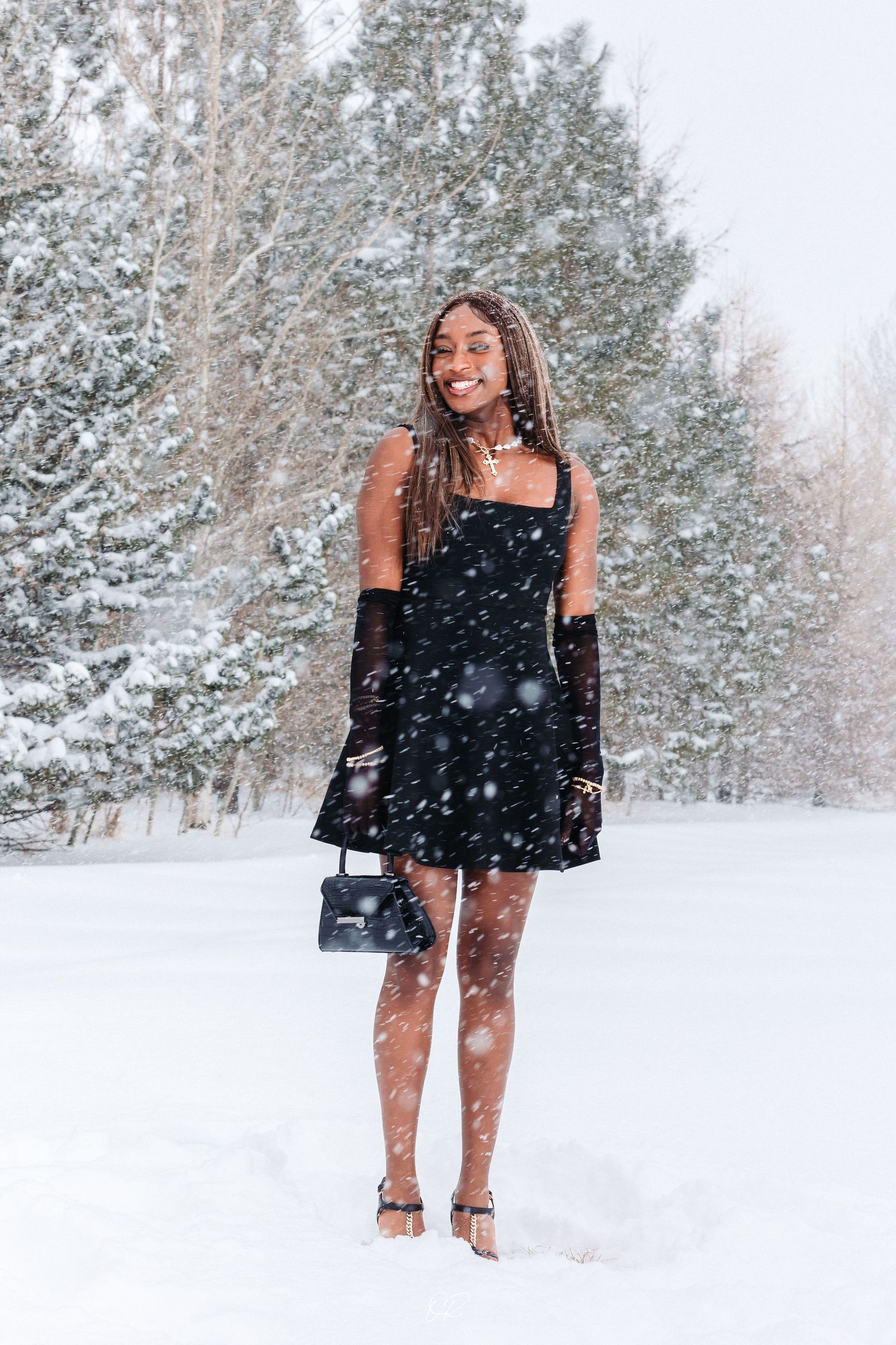 Person in a black dress and gloves poses joyfully in the falling snow with snow-covered trees in the background.
