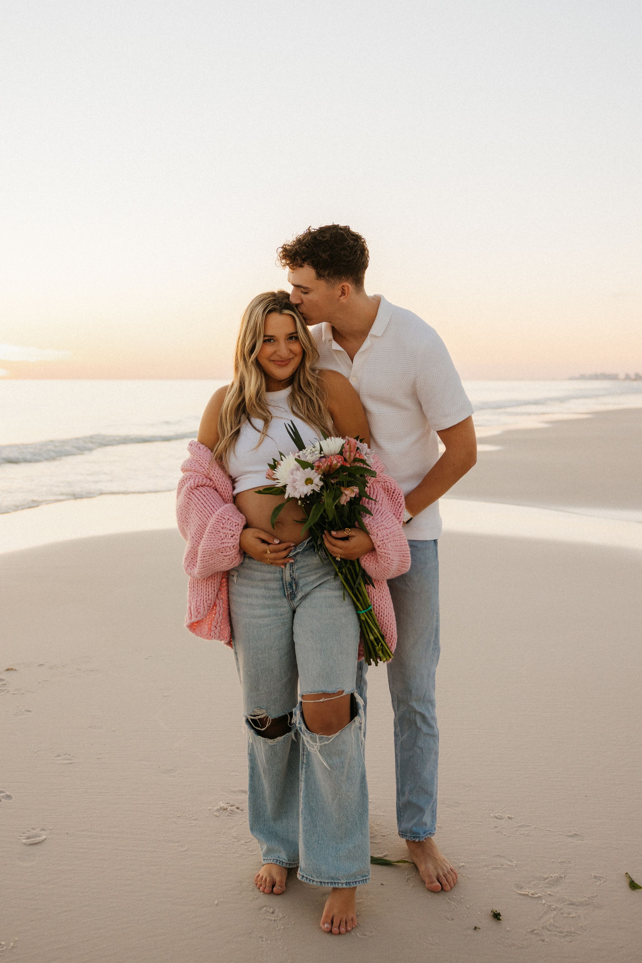 Romantic couple embraces on beach at sunset while holding fresh flowers, wearing casual denim and pink sweater.