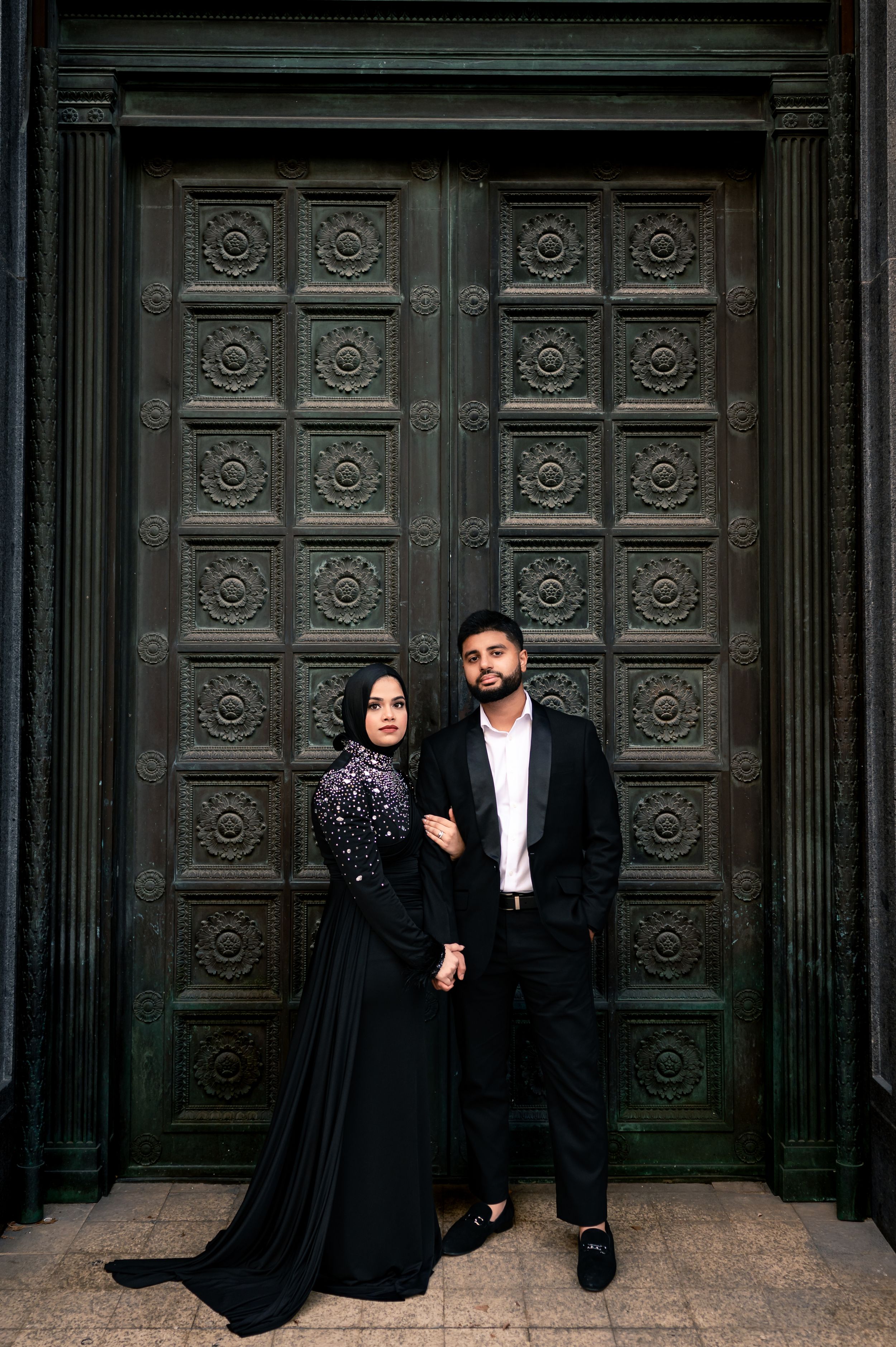 A couple in black formal attire poses dramatically against ornate vintage doors in a romantic photoshoot series.