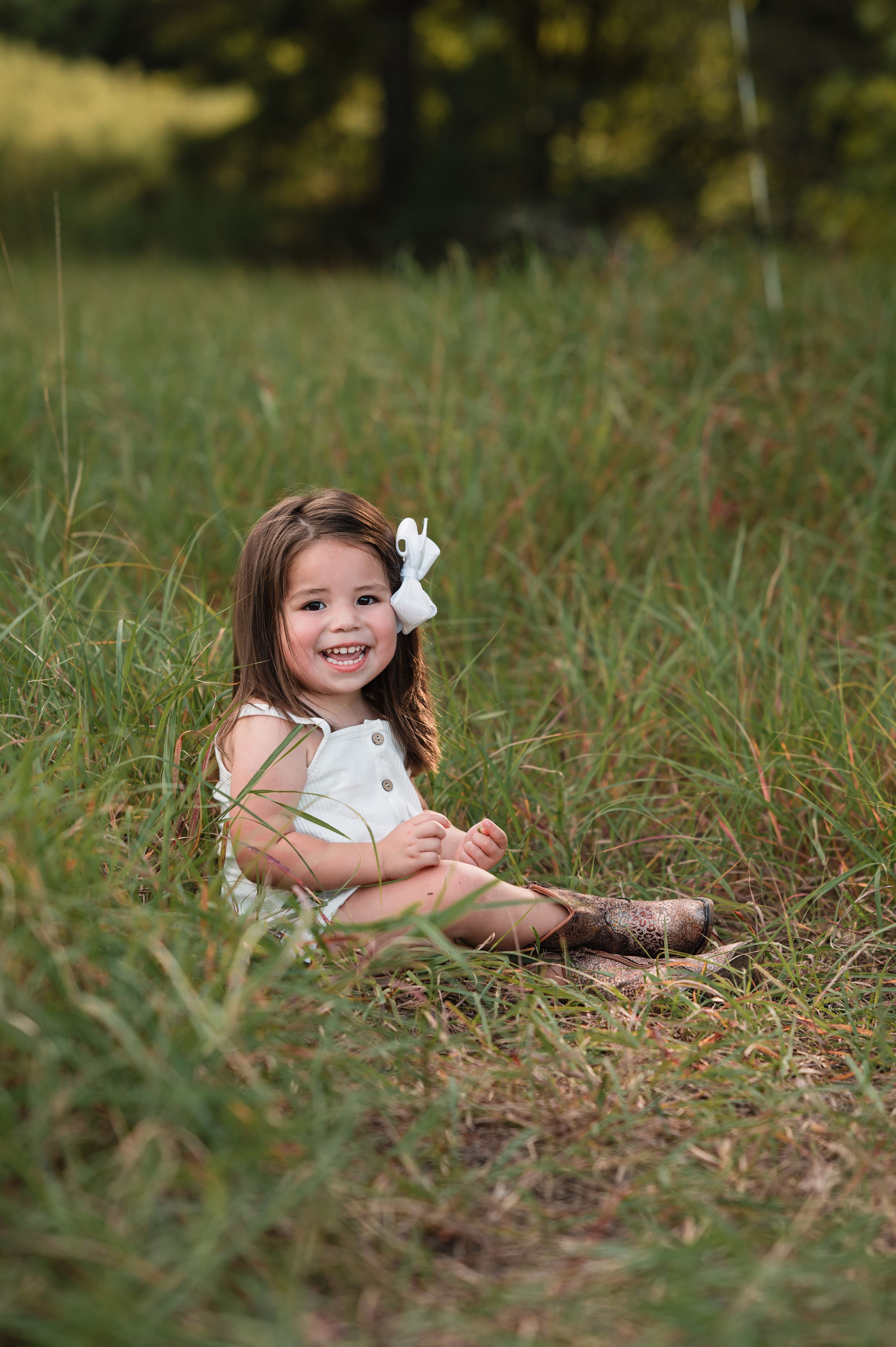 A small child wearing a white dress lies playfully in tall grass during an outdoor photo session.