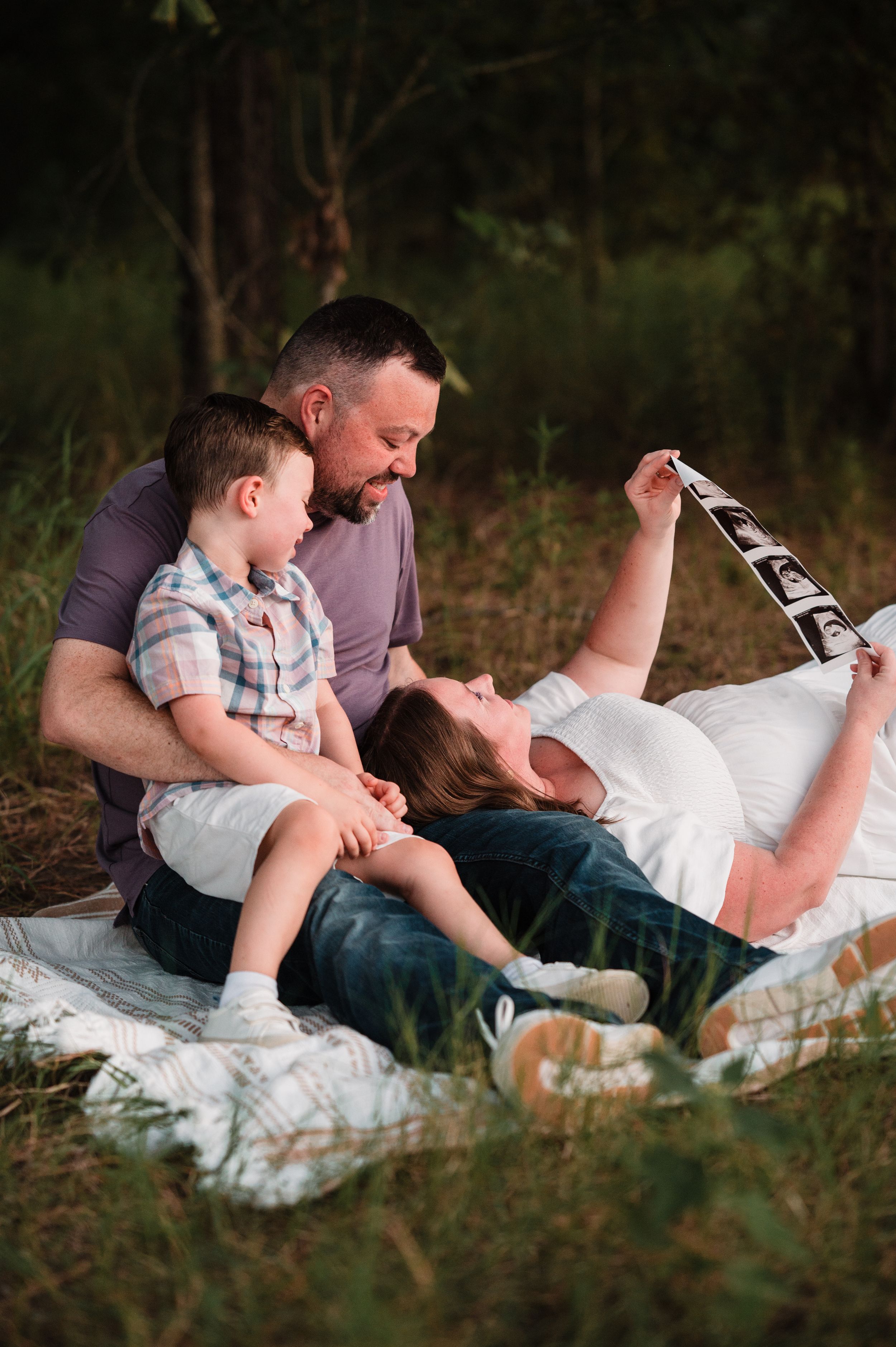 Family lying together on a white blanket outdoors during a casual photo session.