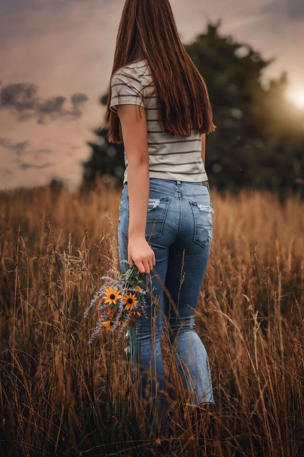 A woman in blue jeans holds sunflowers while standing in a golden autumn field at sunset.