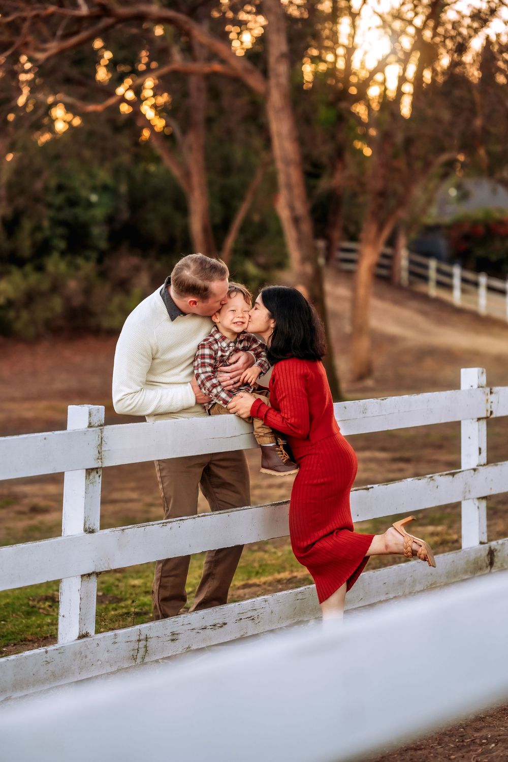 A couple in rustic countryside setting shares romantic moments along white fence at sunset on their engagement shoot.