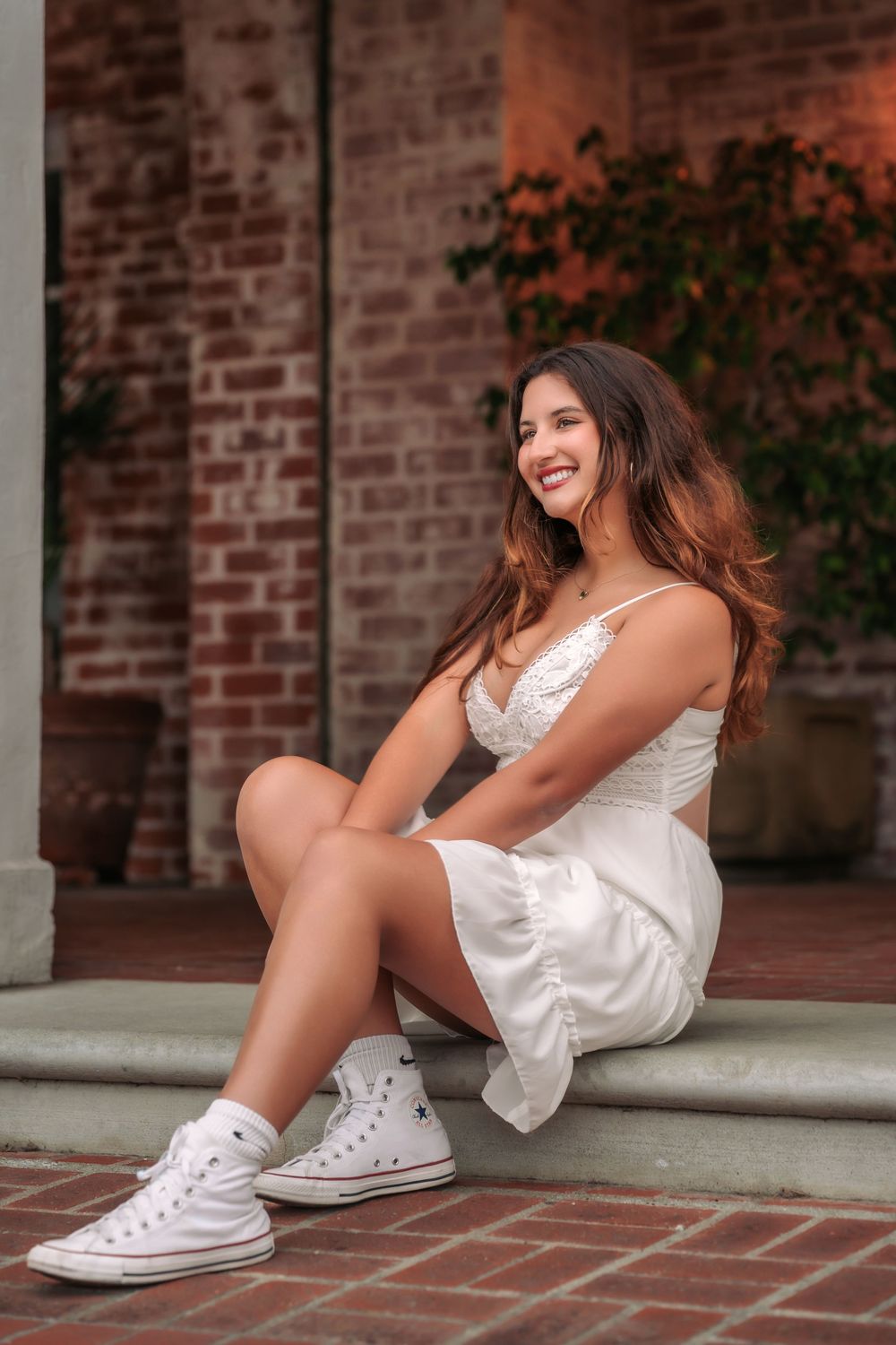 A person in a white dress and white Converse sneakers sits casually on brick steps against a rustic wall.