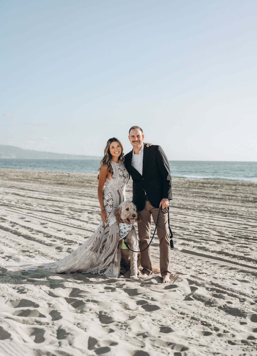 A couple shares a sweet moment while standing together on a sandy beach with ocean waves visible in the background.