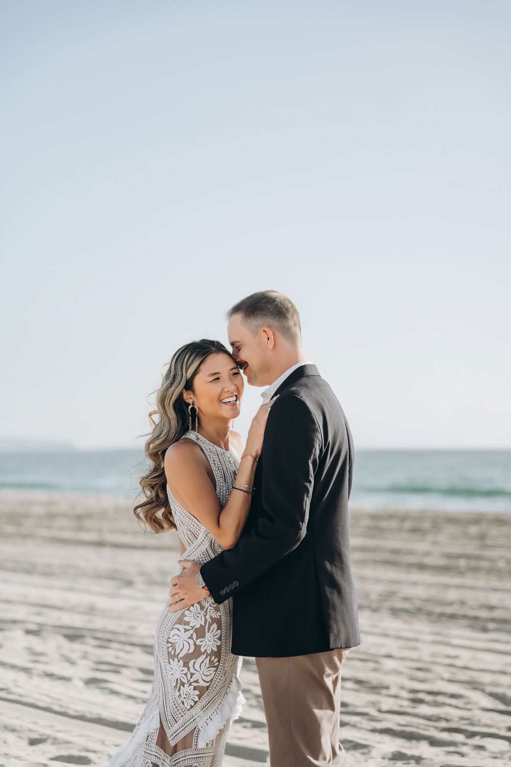 A romantic couple embraces and laughs together on a peaceful beach during a sunny afternoon photoshoot.