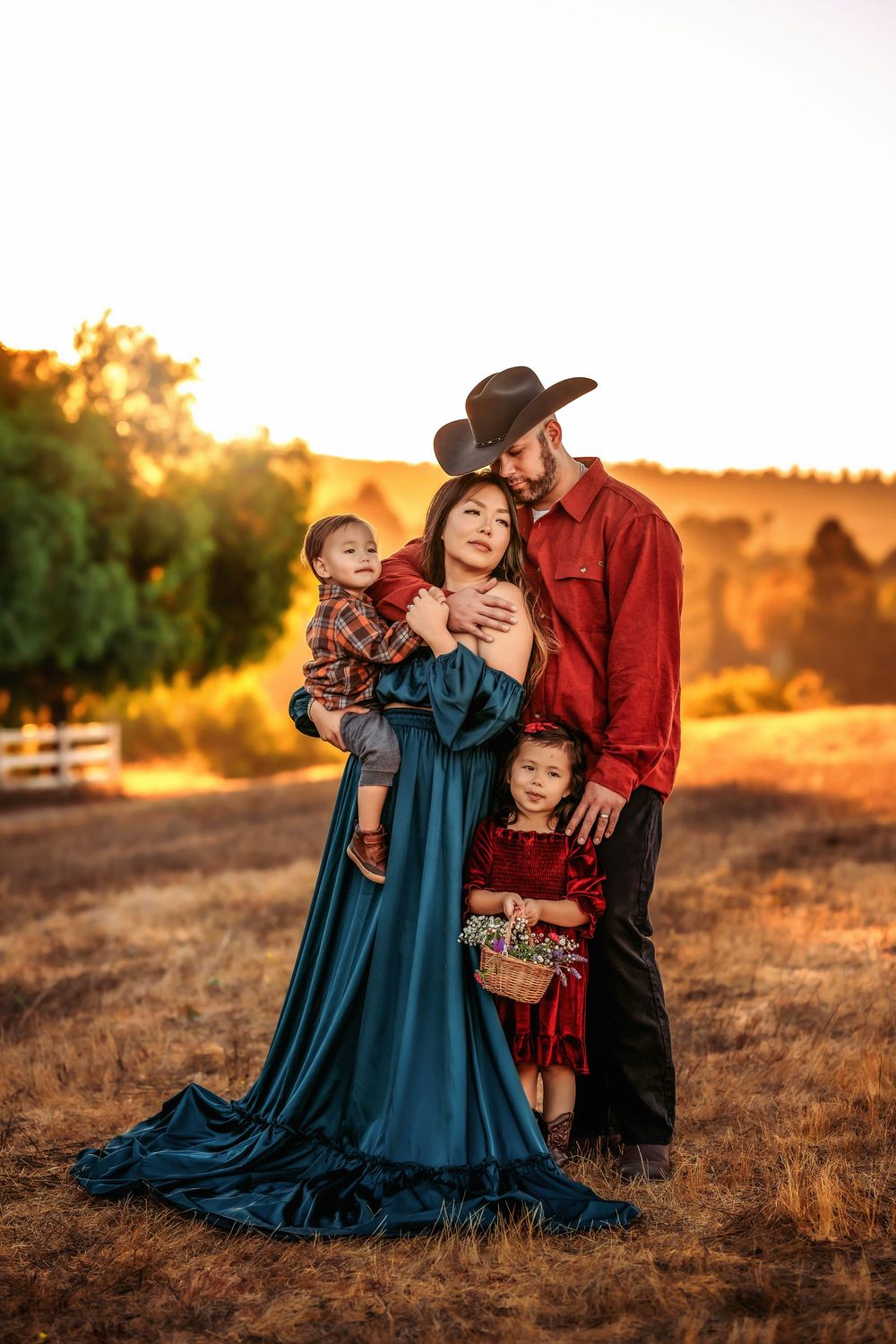 Family wearing western style clothing poses together in a golden field at sunset, with mother in flowing teal dress.