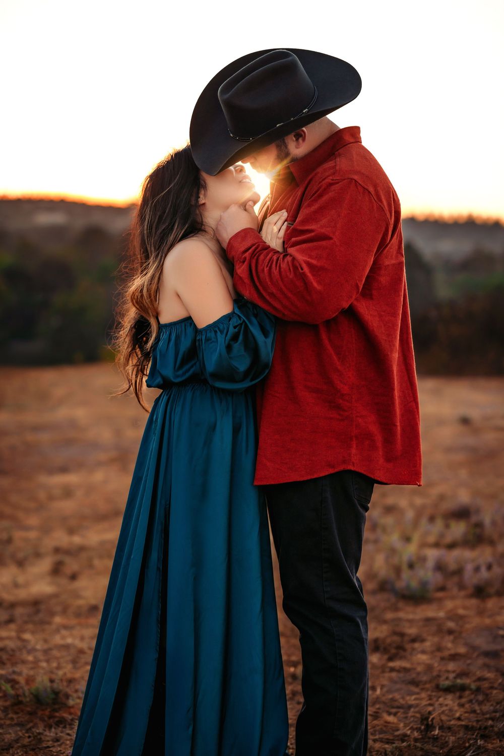 A couple in a red shirt and blue dress share an intimate moment during sunset in a desert landscape.