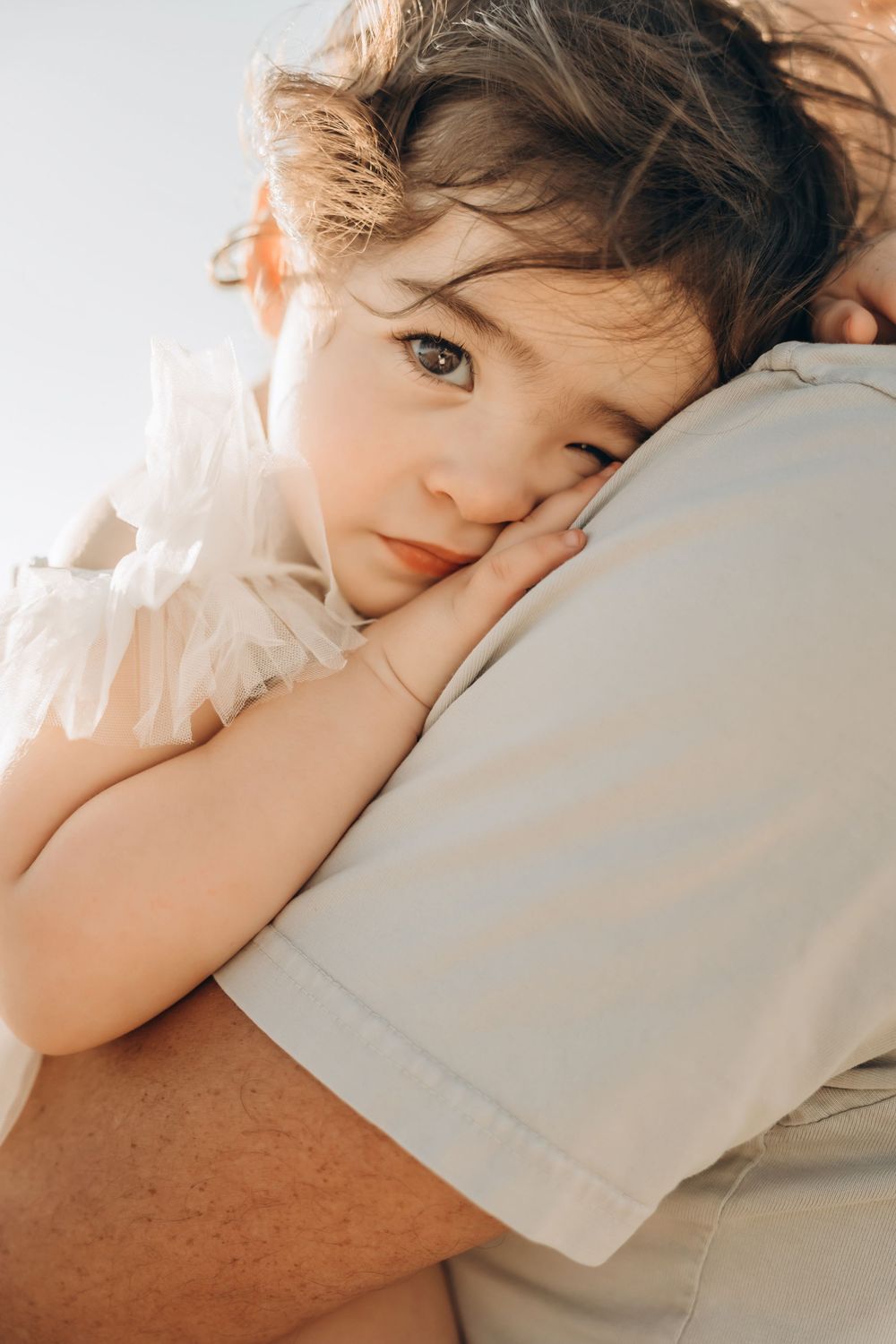 A young child in white clothing rests their head gently against someone's shoulder in soft warm lighting.