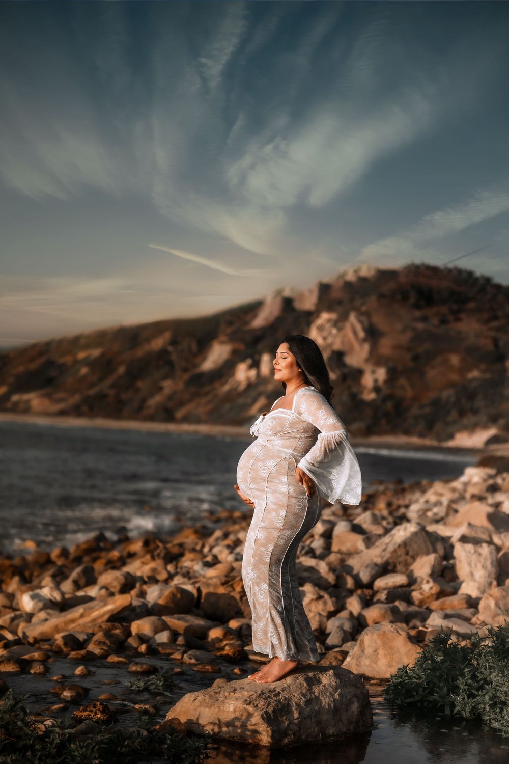 A sunset maternity photoshoot on a rocky beach with dramatic coastal cliffs and warm golden lighting.