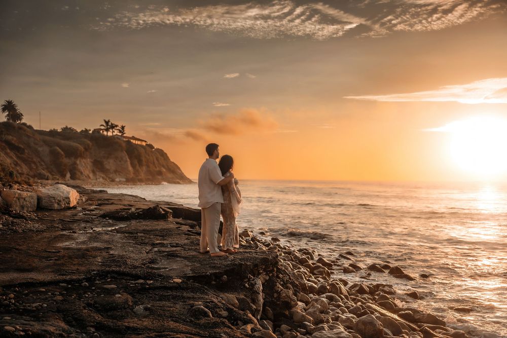 A romantic sunset silhouette scene on a rocky cliff overlooking the Pacific Ocean with golden light illuminating the coast.