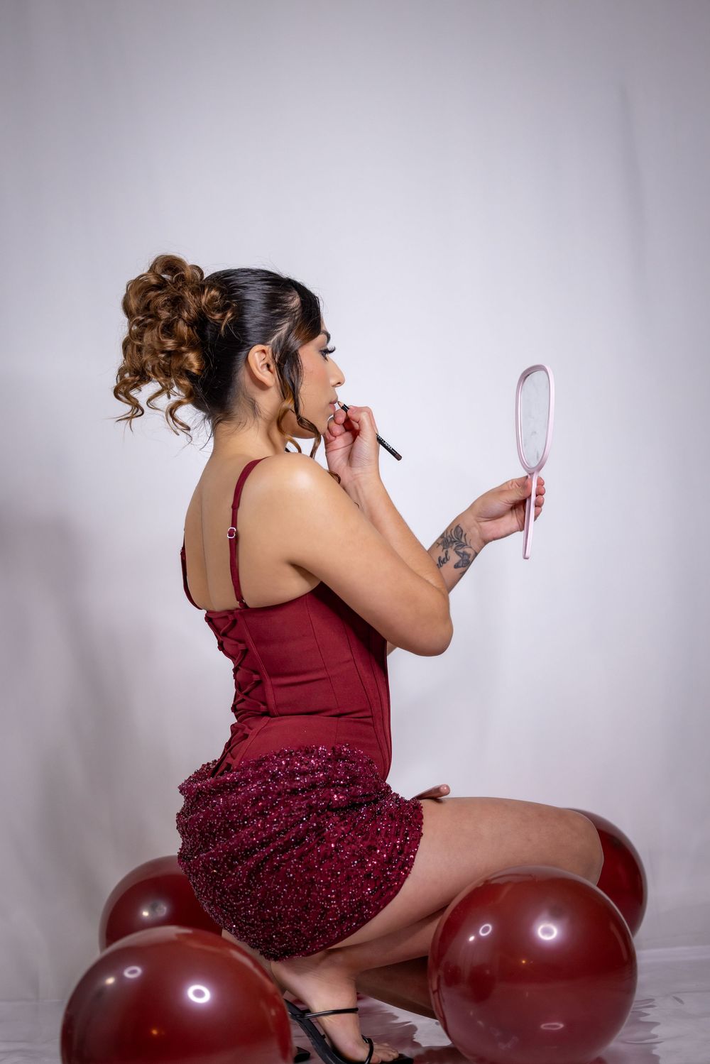 A person in a burgundy dress poses with large red balloon decorations against a white studio backdrop.