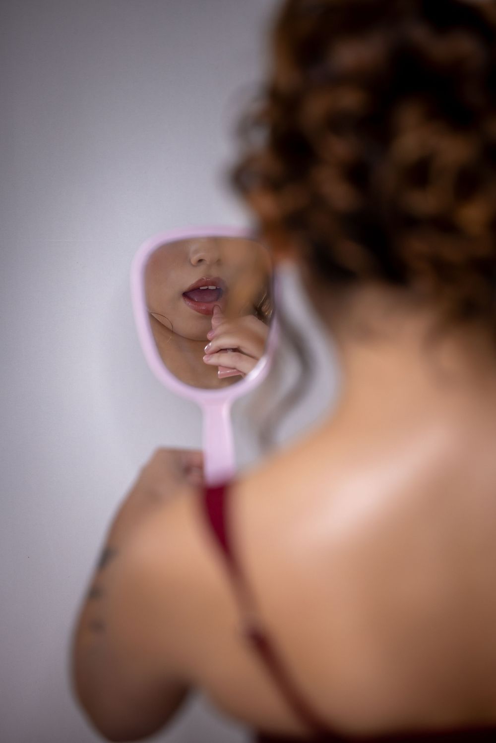 A reflection of red lips and curly hair appears in a small pink handheld mirror against a light background.